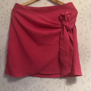 Rosa omlottkjol med rosett - Snygg rosa omlottkjol med en stor rosett på sidan. Kjolen har en elegant omlott-design som ger en fin passform. Perfekt för att lägga till en färgklick i garderoben!