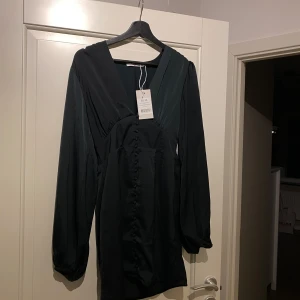 Svart klänning med knappar - Elegant svart klänning med långa ärmar och knappar framtill. Perfekt för en stilren look. Klänningen har en v-ringad design och är tillverkad i ett mjukt material som ger en fin passform.