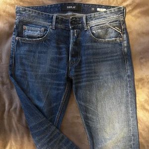 Blå jeans från Replay - Snygga blå jeans från Replay med klassisk femficksdesign och diskreta slitningar. De har en normal passform och är perfekta för en avslappnad stil. Märkesdetaljer syns på fickan och baksidan. Knappen är av där av priset, men går lätt att fixa!