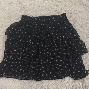 Svart blommig volangkjol - Charmig svart kortkjol med blommigt mönster och volanger. Perfekt för en trendig look med elastisk midja för bekväm passform.