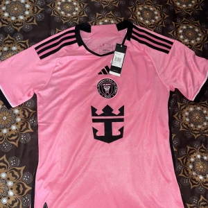Rosa fotbollströja från Adidas - Säljer en rosa fotbollströja från Adidas med svart tryck och detaljer. Tröjan har korta ärmar och klubbmärket för Club Internacional de Fútbol Miami på bröstet. Den är märkt med 'AEROREADY' för optimal komfort. Baksidan har namnet 'Messi' och nummer 10.