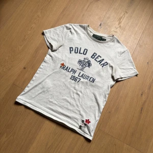 Vit t-shirt med Polo Bear från Ralph Lauren - Snygg vit t-shirt från Ralph Lauren med det ikoniska Polo Bear-motivet och texten 'Ralph Lauren 1967'. T-shirten har korta ärmar och är broderad med blommor för en unik touch. Perfekt för en avslappnad stil.