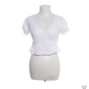 Vit blus med volangdetaljer - Söt vit blus med v-ringning och volangdetaljer. Kortärmad design med en lätt croppad passform som ger en feminin touch. Perfekt för varmare dagar.