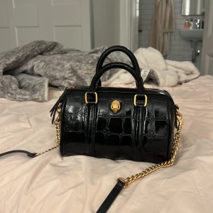 Kurt Geiger väska  - Snygg svart handväska i skinn med krokodilmönster från Kurt Geiger. Väskan har guldfärgade detaljer och en kedja som axelrem. Den har en dragkedja upptill och ett unikt emblem framtill. Köpt under jullovet i London. 