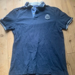 Blå pikétröja med nummerdetalj - Säljer en blå pikétröja med korta ärmar och en broderad logga på bröstet. Tröjan har en krage med knappar och en stor siffra '1' på ärmen. 