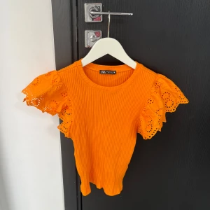 Orange ribbad topp från Zara - Snygg orange ribbad topp från Zara med korta ärmar i spets. Perfekt för att liva upp garderoben med en pop av färg. Den har en rund halsringning och är tillverkad i ett mjukt material.
