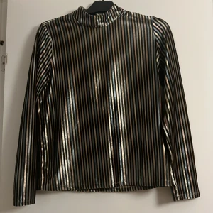 Svart och guld randig långärmad topp - Snygg långärmad topp med svart och guld randigt mönster. Perfekt för en glammig look med sin glittriga finish. Toppen har en hög krage och är tillverkad i ett glänsande material.