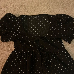 Svart prickig blus - Charmig svart blus med vita prickar och puffiga kortärmar. Blusen har en söt knytning framtill och en lätt rynkad design som ger en feminin touch. Perfekt för en avslappnad och stilfull look. (Man borde ha ett linne under rekommenderar de iaf, hade inte de på bilden) använd fåtal gånger och köparen står för frakten💕💕