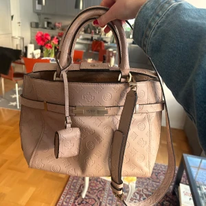 Beige handväska från Guess - Snygg beige handväska från Guess med präglat mönster och praktisk dragkedja. Väskan har både handtag och en avtagbar axelrem för flexibilitet. Perfekt för att bära dina dagliga nödvändigheter med stil.