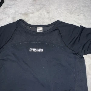 Svart träningströja från Gymshark - Säljer en svart träningströja från Gymshark. Tröjan har korta ärmar och är tillverkad i ett lätt och andningsbart material, perfekt för intensiva träningspass. Gymshark-loggan är tryckt på bröstet. Passar utmärkt för gymmet eller löprundan.