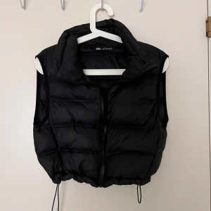 Svart dunväst från Zara - Snygg svart väst från Zara med dragkedja framtill. Västen har en hög krage och justerbar nederkant med dragsko. Material 100% polyester