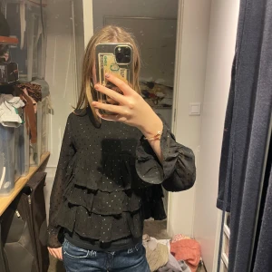 Svart blus med volanger - Säljer en superfin svart blus med volanger och prickigt mönster. Den har långa ärmar och en luftig passform, perfekt för både vardag och fest. Blusen är i mycket bra skick och ger en elegant touch till vilken outfit som helst. Ifrån Gina tricot har haft på mig en gång på Nobel dagen. Säljer för att den inte riktigt är min still. Original pris 299