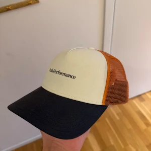 Truckerkeps från Peak Performance - Snygg truckerkeps från Peak Performance med vit front och svart skärm. Sidorna och baksidan är i orange mesh för bra ventilation. Perfekt för sommarens äventyr eller en avslappnad stil. Justerbar passform för att sitta bekvämt hela dagen.