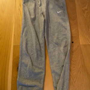 Grå mjukisbyxor från Nike storlek 147-158 cm - Säljer ett par grå mjukisbyxor från Nike i bra skick. De har en normal passform och är perfekta för träning eller chill. Byxorna har en elastisk midja och en liten Nike-logga på sidan. Superbekväma och stilrena för vardagsbruk!