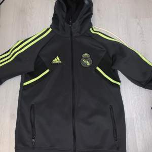 Säljer en svart träningsjacka från Adidas med Real Madrid-logga. Den har neongula detaljer och dragkedja framtill. Perfekt för fotbollsträning eller som en sportig vardagsjacka. Huvan ger extra skydd mot väder och vind.