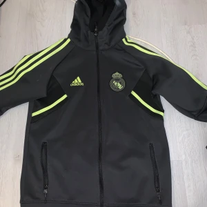 Svart träningsjacka från Adidas - Säljer en svart träningsjacka från Adidas med Real Madrid-logga. Den har neongula detaljer och dragkedja framtill. Perfekt för fotbollsträning eller som en sportig vardagsjacka. Huvan ger extra skydd mot väder och vind.