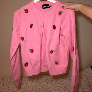 Rosa kofta med jordgubbsmönster från Pieces - Supergullig rosa kofta från Pieces med små jordgubbar broderade över hela. Perfekt för att liva upp garderoben! Den har långa ärmar och knäppning framtill. 🌸🍓
