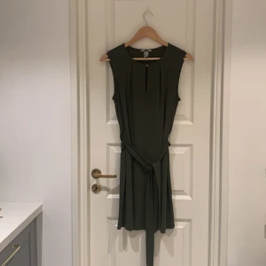 Olivgrön ärmlös klänning med knytband - Säljer en stilren olivgrön klänning i mycket bra skick. Den är ärmlös och har ett knytband i midjan för en smickrande passform. Perfekt för både vardag och festliga tillfällen. Materialet känns mjukt och bekvämt mot huden.