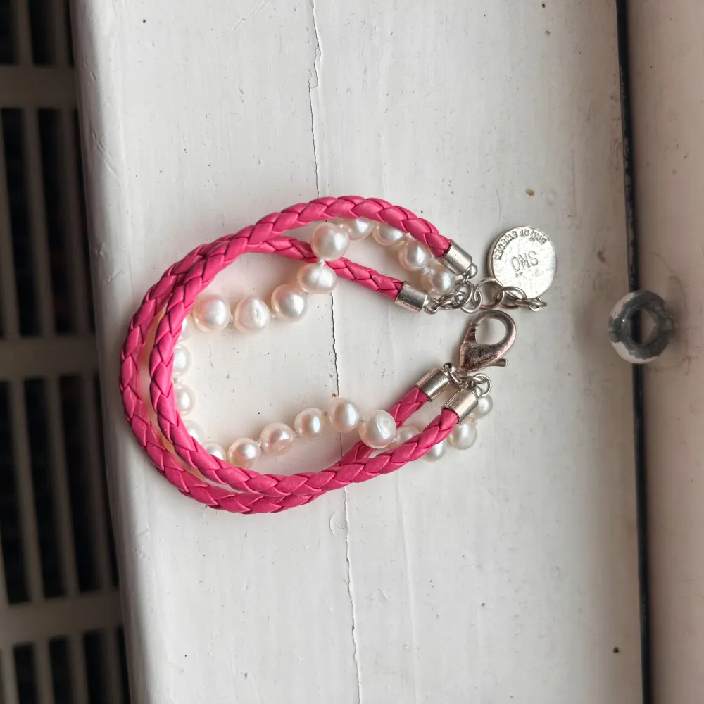 Snyggt armband med en mix av rosa flätat band och vita Sötvattenpärlor . Armbandet har en silverfärgad kedja och ett litet runt hänge. Perfekt för att ge en färgklick till din outfit!. Asusteet.