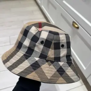 Snygg bucket hat från Burberry i klassiskt rutigt mönster. Hatten har en beige, svart och röd färgkombination som ger en stilren look. Perfekt för både vår och sommar. Den är i mycket bra skick och passar perfekt för en trendig outfit. Bild 3 så ser ni dock ett streck med smusts på hatten, har inte testat rengöra den då jag har tröttnat på den och inge orkat lägga ner tid och enargi. Skulle säga det är mer s än m i storleken på hatten.