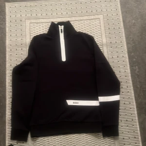 Svart tröja med dragkedja från Boss - Säljer en stilren svart tröja från Boss med vit dragkedja och detaljer. Tröjan har en hög krage och en vit rand på ärmen samt Boss-logga på framsidan. Perfekt för en casual look eller träning.