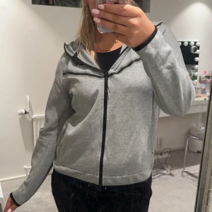 Grå Nike tech fleece - Säljer en snygg och bekväm nike tech fleece. Den har en praktisk ficka på ärmen och en luva, även fickor i sidan. 