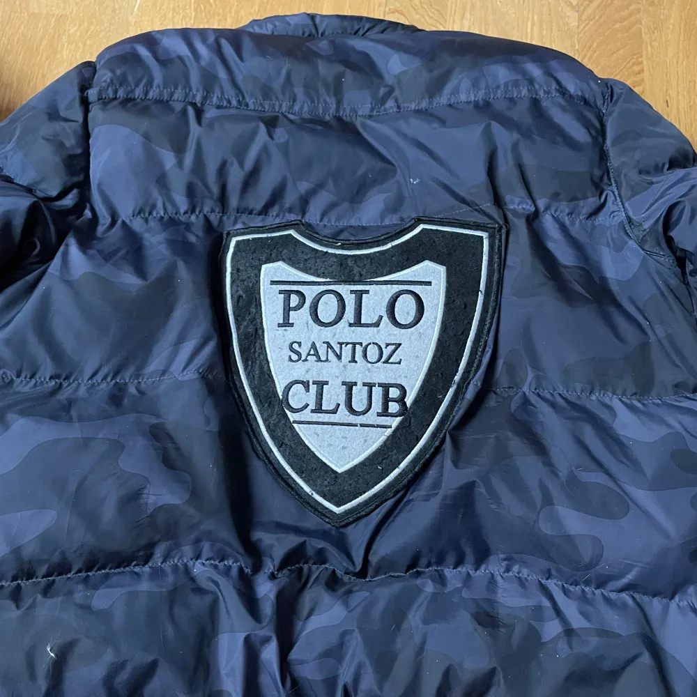 Säljer en snygg svart pufferjacka från Santoz Polo Club. Jackan har ett coolt camouflagemönster och stora logotryck både fram och bak. Den är långärmad med dragkedja och har en röd insida som ger en schysst kontrast. Perfekt för höst och vinter!. Jackor.