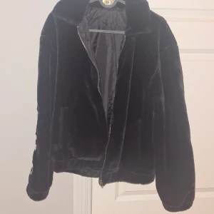 Svart pälsjacka från Sweet SKTBS - Säljer en supermjuk och fluffig svart pälsjacka från Sweet SKTBS. Den har en dragkedja framtill och långa ärmar med texten 'SWEET SKTBS' i vitt på ärmarna. Perfekt för att hålla sig varm och stilren under kyliga dagar. Passar både till vardags och fest!