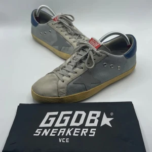 Grå GGDB sneakers med blå detaljer - Säljer ett par GGDB sneakers i grått med blå detaljer och en stjärna på sidan. Skorna har en vintage look med snörning och en slitstark gummisula. Perfekta för en avslappnad stil.