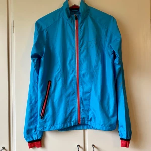 Blå löparjacka med rosa detaljer - Säljer en snygg blå löparjacka med rosa dragkedjor och muddar. Perfekt för blåsiga dagar och har en sportig look. Jackan är långärmad och har en normal passform. Den är i bra skick och redo för nya äventyr!