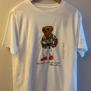 T-shirt Ralph Lauren - Säljer en snygg vit t-shirt från Ralph Lauren med ett coolt björntryck på framsidan. Storleken är XL barn, men passar S herr. T-shirten är i nytt skick och gjord av mjuk bomull. Aldrig använd.