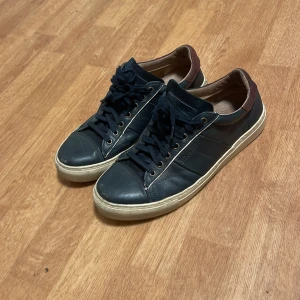 Mörkblå sneakers  - Säljer ett par Mörkblå sneakers i skinn med bruna detaljer vid hälen. De har en klassisk design med snörning och en vit sula som ger en snygg kontrast. Perfekta för vardagsbruk och i bra skick. Passar till det mesta i garderoben!