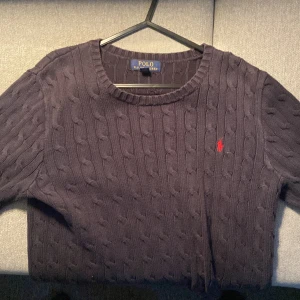 Mörkblå stickad tröja från Polo Ralph Lauren - Säljer en snygg mörkblå stickad tröja från Polo Ralph Lauren. Den har ett klassiskt kabelstickat mönster och en liten röd logga på bröstet. Perfekt för höst och vinter, med långa ärmar och rund halsringning. Passar både till vardags och lite finare tillfällen.