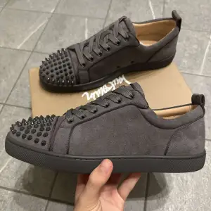 Snygga grå sneakers i mocka med coola nitar på tån och den ikoniska röda sulan. Perfekta för att ge din outfit en edgy touch. Skorna har snörning och är i mycket bra skick. Passar både till vardags och fest! Storlek 44