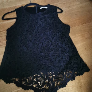 Svart spetslinne från Cutey - Snyggt svart spetslinne från Cutey i storlek d. Det är ärmlöst och har ett vackert broderat mönster över hela framsidan. Perfekt för en festlig kväll eller en stilren vardagslook. Linnet har en elegant avslutning med spetsdetaljer längst ner.