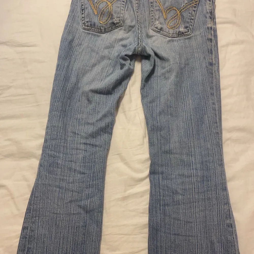 Säljer ett par snygga blå bootcut jeans från Bebe i storlek 26. De har en klassisk femficksdesign och är dekorerade med guldfärgade broderier på bakfickorna. Perfekta för en avslappnad vardagslook eller en utekväll. Skicket är bra och de är tillverkade i USA.. Jeans & Byxor.