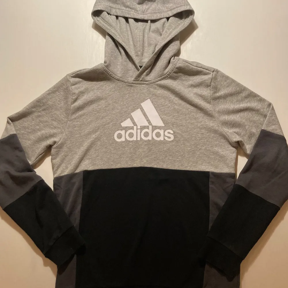 Säljer en snygg och bekväm grå och svart hoodie från Adidas. Den har en stor logga på bröstet och är perfekt för både träning och vardag. Hoodien har långa ärmar och en klassisk passform. Perfekt för höst och vår!. Tröjor & Koftor.