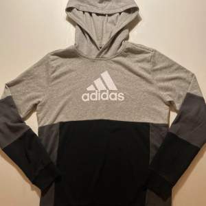 Säljer en snygg och bekväm grå och svart hoodie från Adidas. Den har en stor logga på bröstet och är perfekt för både träning och vardag. Hoodien har långa ärmar och en klassisk passform. Perfekt för höst och vår!