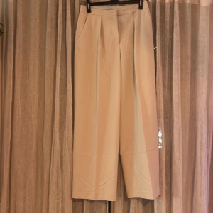 Beige kostymbyxor från H&M - Säljer ett par superfina beige kostymbyxor från H&M i storlek 42, ganska små i storleken. Byxorna har en högmidjad passform och är tillverkade i ett mjukt och bekvämt material. Perfekta för både kontoret och festliga tillfällen. De har en klassisk design med raka ben och diskreta fickor bak. Knappt använda. 
