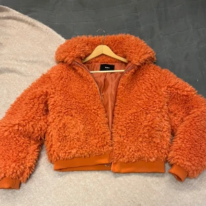 Orange teddyjacka - Säljer en oanvänd supermysig orange teddyjacka som är perfekt för höst och vinter. Jackan har en dragkedja framtill och är i mycket bra skick. Den är fluffig och varm, perfekt när det är kallt ute. 