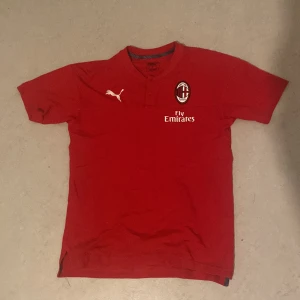 Ac Milan fotbollströja  - Säljer en röd Ac Milan fotbollströja från Puma med korta ärmar. Tröjan har en snygg design med klubbmärke och Fly Emirates-logga på framsidan. Perfekt för fotbollsfans som vill visa sitt stöd. Tröjan är i bra skick och redo för nya äventyr på planen eller i soffan.