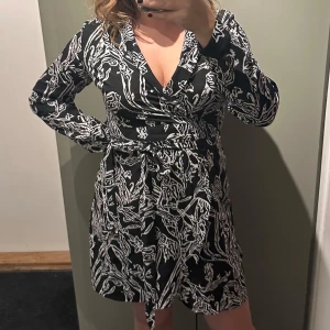 Svart och vit omlottklänning från Neverfullydressed  - Snygg svart och vit omlottklänning från Neverfullydressed i storlek M/L. Klänningen har ett abstrakt mönster och långa ärmar, perfekt för både vardag och fest. Den är tillverkad i ett mjukt och bekvämt material som ger en smickrande passform.