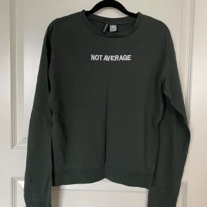 Mörkgrön tröja från Divided - Säljer en mörkgrön tröja från Divided med texten 'NOT AVERAGE' tryckt på framsidan. Tröjan har långa ärmar och en rund halsringning. Perfekt för en avslappnad stil och passar bra till både jeans och kjol. Skön och enkel att bära!