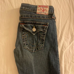 Blå jeans från True Religion - Säljer ett par snygga blå jeans från True Religion i bra skick. De har en bootcut-stil och är lågmidjade med coola detaljer som knappar och broderade bakfickor.  Innerbenslängd: 79 cm midjemått:37cm