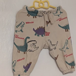 Beige barnbyxor med dinosauriemönster, Strl 74 - Supergulliga beige barnbyxor från H&M i storlek 74. Byxorna har ett roligt dinosauriemönster i färgerna blå, grön och röd med texten 'Little Brosaurus'. De är gjorda av 100% bomull och har en bekväm resårmidja med snörning. Perfekta för små äventyrare! postar varje söndag och fredag 