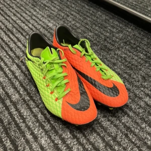 Nike fotbollsskor i neonfärger - Säljer ett par coola Nike fotbollsskor i neonorange och neongrönt. Skorna har en snygg svart Nike-logga och är perfekta för fotbollsträning. De har snörning och en mönstrad yta för extra grepp på planen. Använda men i bra skick, redo för nya äventyr på fotbollsplanen! ⚽