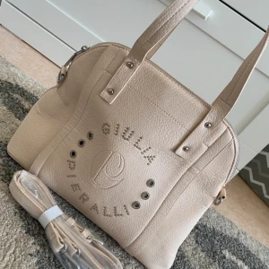 Beige handväska från Giulia Pieralli - Snygg beige handväska från Giulia Pieralli i skinn. Väskan har ett stilrent mönster med nitar och logotyp på framsidan. Insidan är fodrad med ett mönstrat tyg och har flera fack för bra förvaring. Perfekt för både vardag och fest!