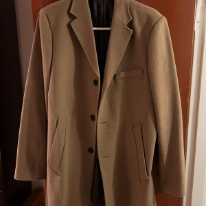 Beige kappa från Bläck - Säljer en stilren beige kappa från Bläck i mycket bra skick. Den har en klassisk design med knappar framtill och fickor på sidorna. Perfekt för höst och vår när du vill se extra snygg ut. Passar både till vardags med en hoodie under och även till lite finare tillfällen.