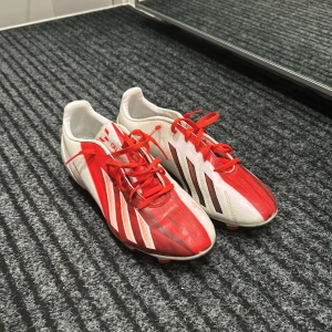 Rödvita fotbollsskor från Adidas - Säljer ett par snygga rödvita Adidas fotbollsskor i mycket bra skick. Skorna har en cool design med röda och vita ränder samt snörning. Perfekta för fotbollsträning eller match. De har en robust sula med röda dobbar för bra grepp på planen.