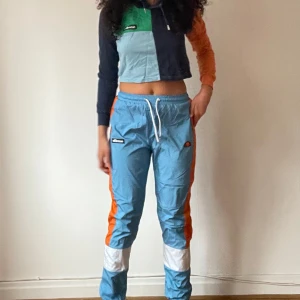 Flerfärgade träningsbyxor från Ellesse - Säljer ett par coola träningsbyxor från Ellesse i blått, orange och vitt. Byxorna har en loose passform med snörning i midjan och är perfekta för träning eller en avslappnad stil. De har också en liten logga på benet. Perfekta för dig som vill sticka ut!